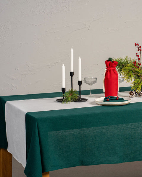 The menique linen tablecloth in dark green color, decorating a Christmas table. there are natural elements, such as pine cones and pine branches on the table top. There is also a wine bottle in a storage bag and candles.