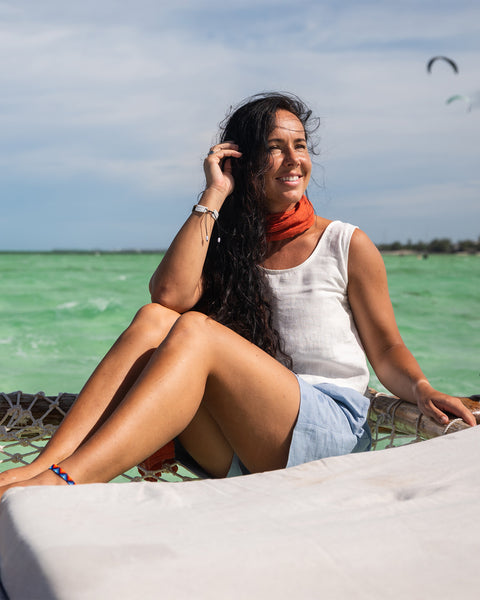 A woman sits in the sun, wearing a white tank top, blue shorts, and an orange scarf, against a backdrop of a bright sky and turquoise water.