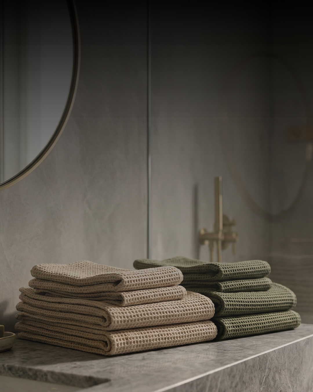 A close-up lifestyle shot of a modern bathroom vanity. Resting on a grey marble countertop are two neatly folded stacks of waffle-weave towels; the stack on the left is light beige, and the stack on the right is olive green. The background features a large, frameless mirror reflecting a gold or brass shower fixture, and the edge of a round, brass-framed mirror is visible on the upper left wall.