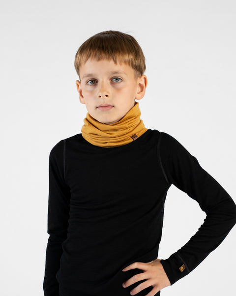 A young boy stands facing forward with one hand on his hip against a white background. He is wearing a black long-sleeved base layer top, black leggings, and a mustard yellow neck gaiter bunched around his neck, featuring a small brown logo tag.