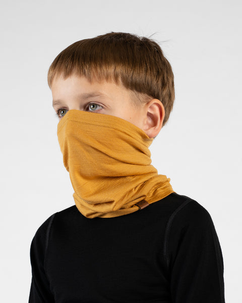 A close-up portrait of a young boy looking off to his left in a three-quarter profile against a white background. He is wearing a black long-sleeved top and a mustard yellow neck gaiter pulled up over his nose and mouth, showing how it bunches comfortably around his neck.