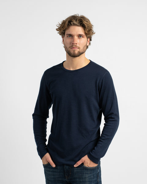A studio portrait of a man standing with his hands loosely resting in his front pockets, wearing a dark navy long-sleeve top and jeans.
