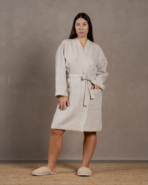The menique women's linen bathrobe in natural color, L-XL size. The woman is standing in front of a gray wall and has a hand in the pocket.