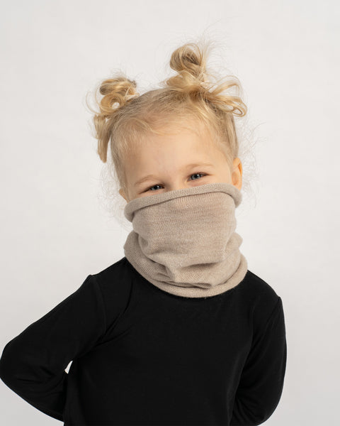 Child playfully covering the lower part of her face with a beige knit neck warmer while wearing a black outfit.