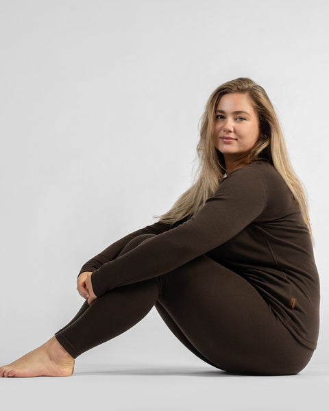 A woman sitting sideways on the floor with knees bent and arms wrapped around them, wearing a dark brown thermal knit set.