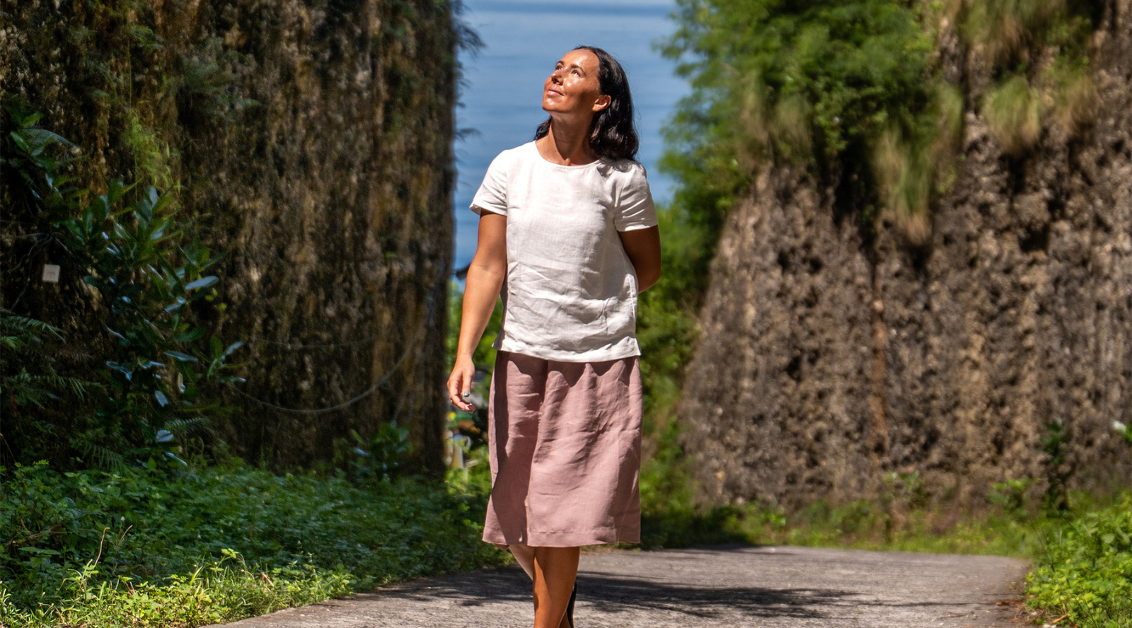 A woman walking along a path between steep rock walls towards a view of the water, wearing a white short-sleeved linen top and a dusty pink linen skirt, looking up towards the sunlight.
