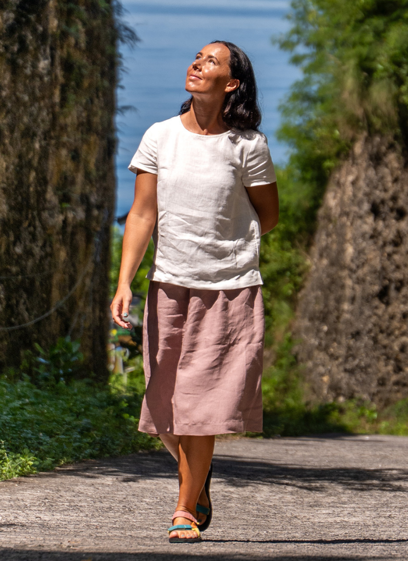 A woman walking along a path between steep rock walls towards a view of the water, wearing a white short-sleeved linen top and a dusty pink linen skirt, looking up towards the sunlight.
