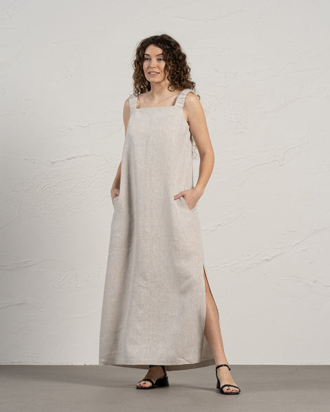 A light-skinned woman with long, dark curly hair stands facing the camera with a gentle smile. She is wearing a sleeveless, menique maxi dress with a square neckline and side slits, and black sandals. Her hands are in the dress pockets, and she stands on a light gray floor against a textured off-white wall.