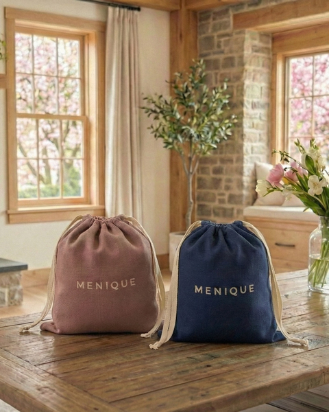 Two Menique branded drawstring gift bags in blush and navy placed on a wooden table in a warm, sunlit living room, showcasing premium packaging for merino wool gifts.