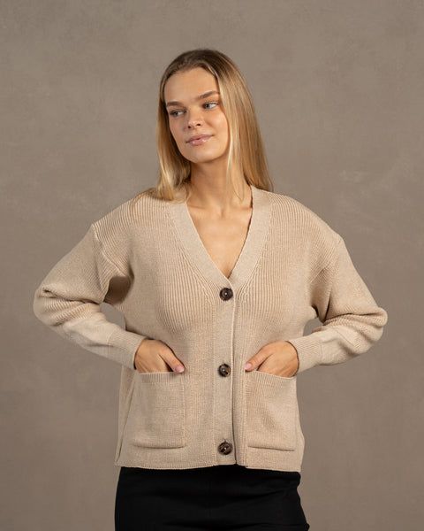 A woman, looking to the side, wearing a beige merino wool knit cardigan. She has both her hands in the front pockets.