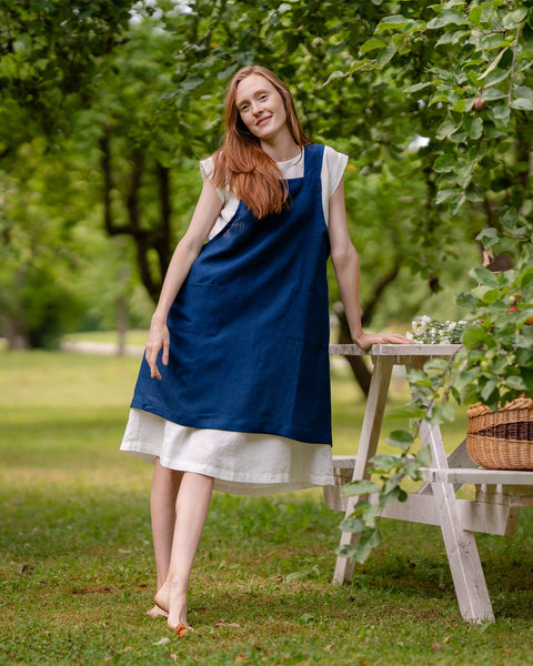 A fair-skinned woman with long reddish hair stands barefoot in a sunny garden wearing a white linen dress under a blue menique linen apron. She lean against a small white table with flowers.