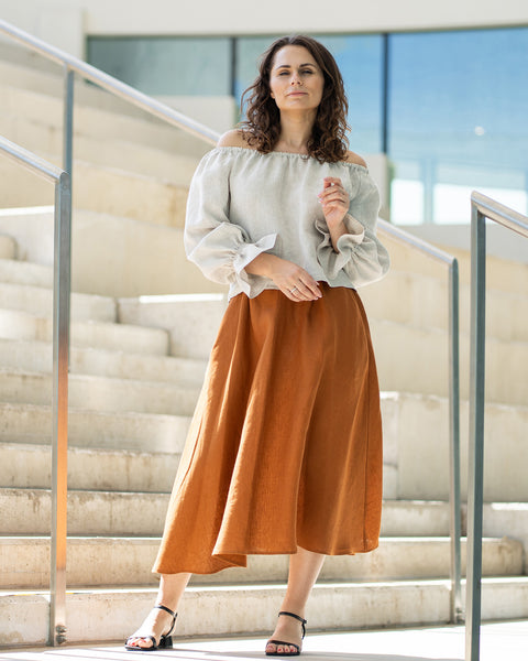 A woman with brown hair, wearing a light beige off-the-shoulder top with puff sleeves and a long rust-brown skirt, poses on concrete stairs.
