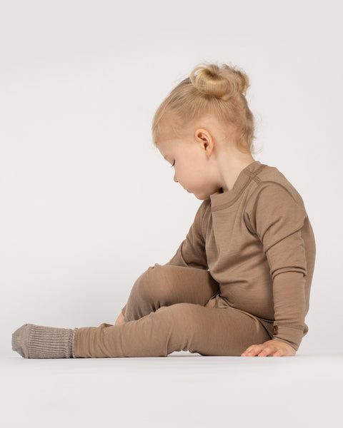 A toddler with blonde hair styled in two small buns sits facing sideways against a plain white background, looking thoughtfully down at the floor. She is wearing a cozy taupe long-sleeve thermal top paired with matching leggings and grey knit socks.