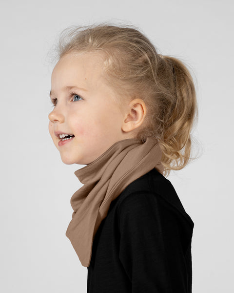 A studio portrait of a young blonde girl looking upwards and smiling slightly. She is seen in profile from the shoulders up. She is wearing a light beige bandana tied around her neck, over a solid black long-sleeved shirt. The background is a plain light grey.