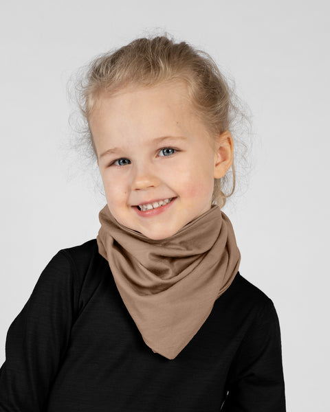 A studio portrait of a young blonde girl looking directly at the camera with a big smile. She is wearing a light beige bandana tied around her neck with the point facing down, over a solid black long-sleeved shirt. The background is a plain light grey.