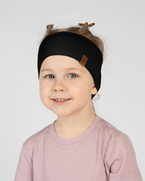A close-up studio portrait of a smiling young child looking directly at the camera against a plain light grey background. She is wearing a light dusty pink short-sleeved t-shirt and a wide black headband with a small brown logo tag on the side, showing ribbon tie details at the top.