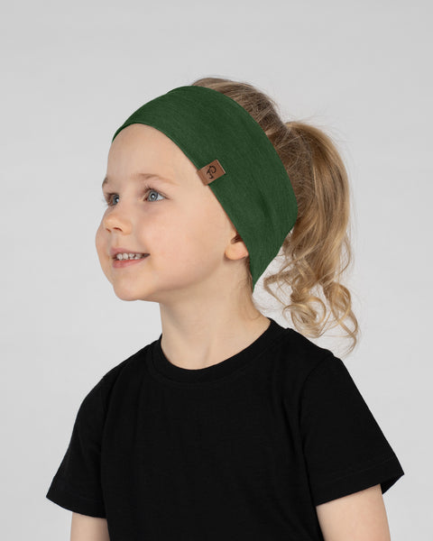 A close-up studio portrait of a smiling young child looking up and to the left against a plain light grey background. She is wearing a black short-sleeved t-shirt and a wide dark green headband with a small brown logo tag on the side, her hair pulled back in a ponytail.