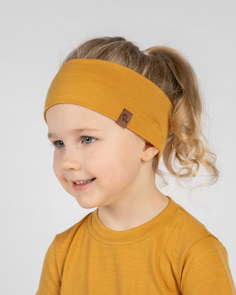 A studio portrait of a smiling young child looking off to the left against a plain light grey background. She is wearing a mustard yellow short-sleeved t-shirt and a matching wide mustard yellow headband with a small brown logo tag on the side, her hair pulled back in a ponytail.