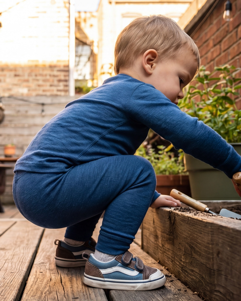 A young boy crouches beside a wooden raised garden bed on an outdoor patio, using a small hand trowel to dig in the dirt. He is wearing a comfortable blue long-sleeve top, matching leggings, and blue and brown Velcro sneakers.