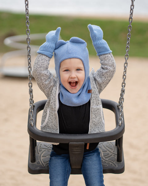 A young child with blue eyes and an open-mouthed expression of excitement, is in a playground swing. They are wearing a light blue balaclava with what look like bunny ears, light blue mittens, a grey speckled zip-up hoodie, and blue jeans, with their arms raised. The background shows a sandy area and some green grass.