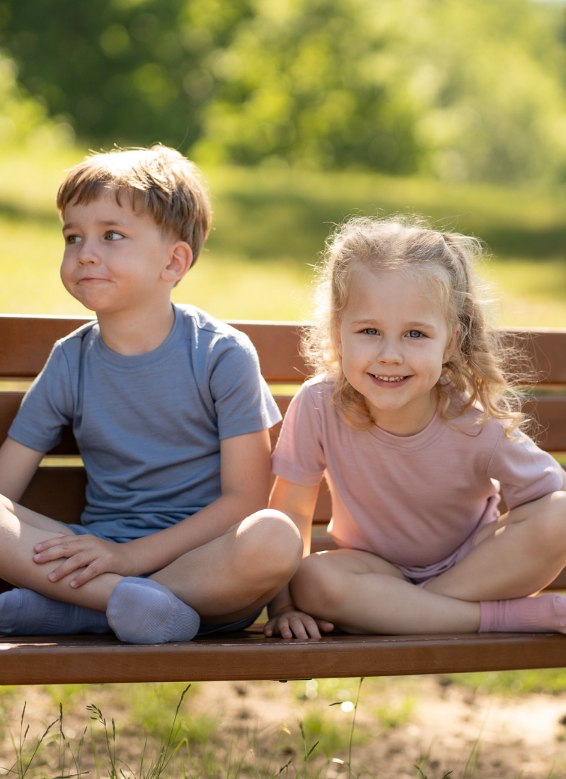 A young boy and girl sitting cross-legged on a wooden park bench outdoors. The boy wears a dusty blue short-sleeve top and shorts set, and the girl wears a dusty pink short-sleeve top and shorts set.