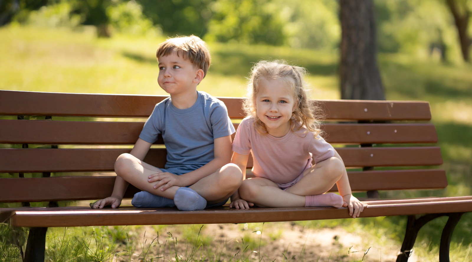 A young boy and girl sitting cross-legged on a wooden park bench outdoors. The boy wears a dusty blue short-sleeve top and shorts set, and the girl wears a dusty pink short-sleeve top and shorts set.