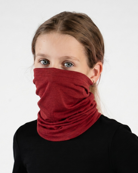A close-up studio portrait of a young girl looking directly at the camera against a white background. She is wearing a black long-sleeved top and a deep red royal cherry neck gaiter pulled up to cover her nose and mouth, showing its long length bunched around her neck.