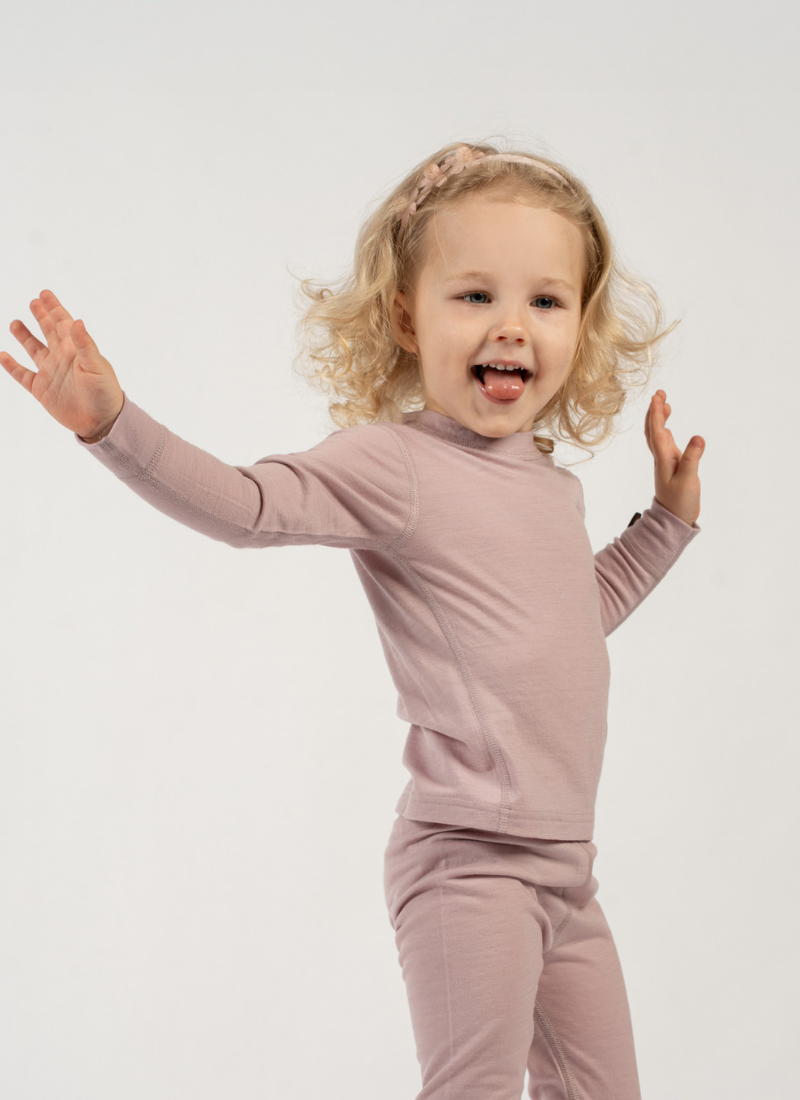 A young girl with curly blonde hair and a small headband stands playfully against a plain white background. She has her arms thrown wide open with an excited expression and her tongue slightly out, wearing a dusty mauve long-sleeve top and matching bottoms.
