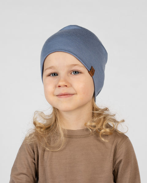 A close-up studio portrait of a young blonde girl with a soft smile looking directly at the camera against a plain light grey background. She is wearing a brown long-sleeved top and a dusty blue beanie featuring a small brown logo tag.