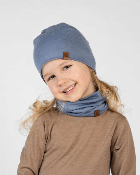 A close-up studio portrait of a smiling young girl tilting her head and looking directly at the camera against a plain light grey background. She is wearing a brown long-sleeved top, a dusty blue beanie, and a matching dusty blue neck gaiter bunched around her neck. Both accessories feature a small brown logo tag.
