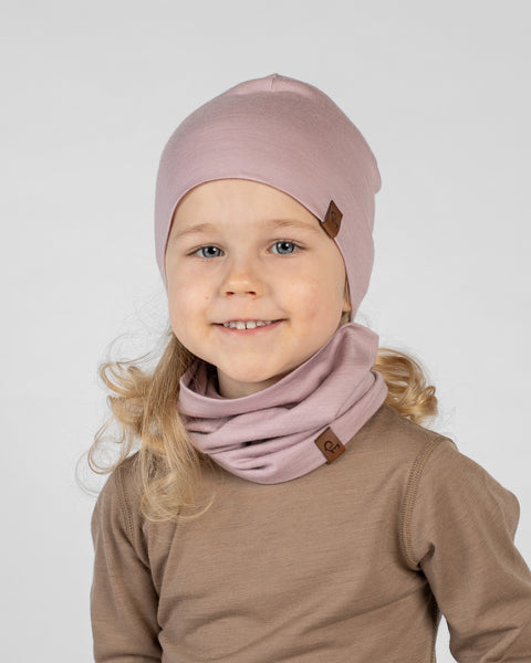A close-up studio portrait of a smiling young girl looking directly at the camera against a plain light grey background. She is wearing a brown long-sleeved top, a dusty pink beanie, and a matching dusty pink neck gaiter bunched around her neck. Both accessories feature a small brown logo tag.