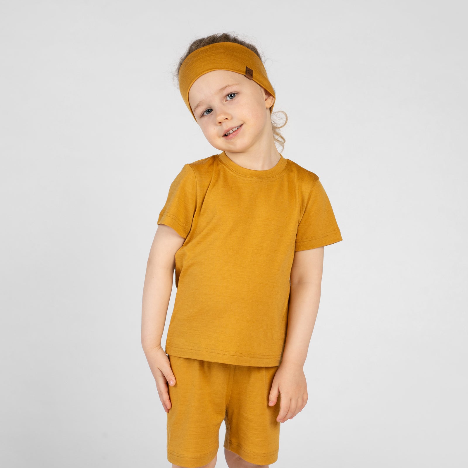 A close-up side profile view of a young child's torso and upper legs. The child is wearing a mustard yellow merino short-sleeve t-shirt and matching mustard yellow merino shorts. The child's hand is slightly lifting the hem of the t-shirt to show the elastic waistband and drawstring of the shorts.