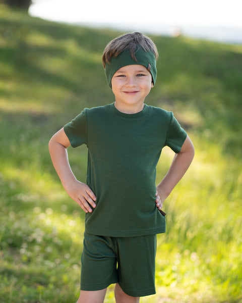 Young boy smiling outdoors wearing a dark green Merino wool T-shirt with a matching Merino wool headband. The soft, breathable outfit and accessory are showcased in a sunny natural setting, offering comfortable everyday wear for active kids.