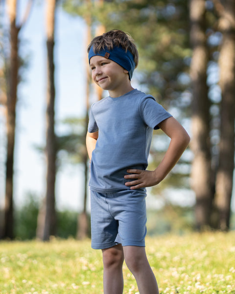 Young boy standing in a sunny park wearing a dusty blue Merino wool T-shirt, matching shorts, and a denim Merino wool headband. The soft, breathable outfit is showcased in a natural outdoor setting, providing comfortable everyday wear for active kids.