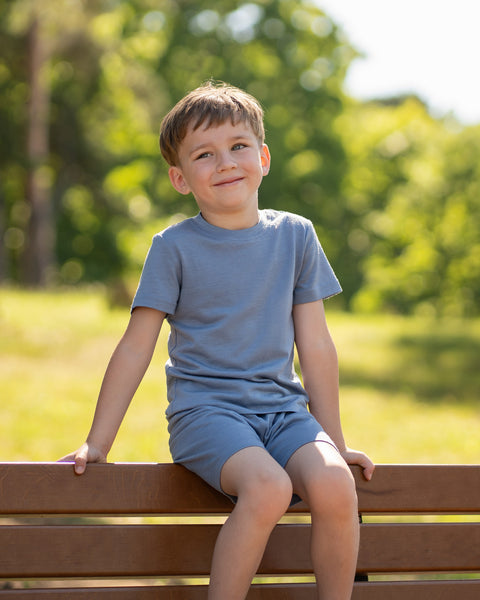 Young boy sitting on a wooden park bench wearing a dusty blue Merino wool T-shirt and matching shorts. The soft, breathable kids' outfit is showcased in a sunny outdoor setting, ideal for everyday comfort and warm-weather adventures.