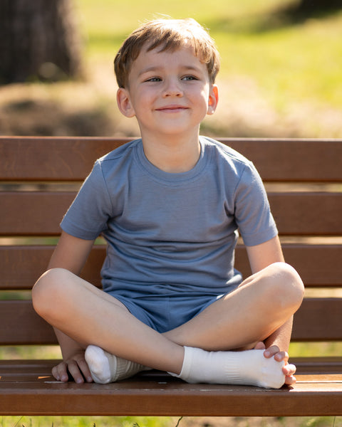 Young boy sitting cross-legged on a wooden park bench wearing a dusty blue Merino wool T-shirt and matching shorts. The soft, breathable kids' outfit is showcased in a sunny outdoor setting, ideal for everyday comfort and warm-weather adventures.