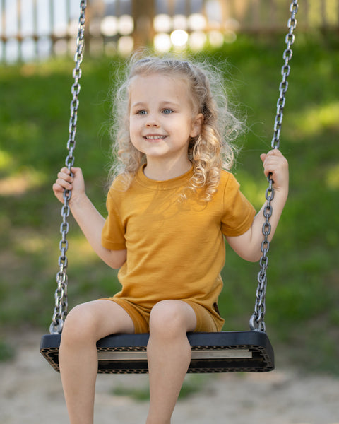 Young girl sitting on a playground swing wearing a yellow Merino wool T-shirt and matching shorts. The lightweight, breathable kids' outfit is showcased in a sunny outdoor setting, offering soft comfort for active play and everyday adventures.