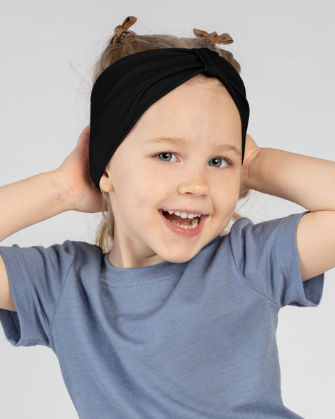 A close-up studio portrait of a young girl smiling brightly at the camera with her mouth open. She is wearing a short-sleeved blue top and a black ribbon headband. She has her hands behind her head against a plain light grey background.