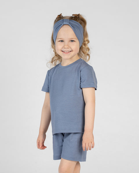 A young girl stands smiling at the camera in a studio setting against a light grey background. She wears a matching short-sleeved top and shorts set in dusty blue, paired with a matching dusty blue ribbon headband in her hair.