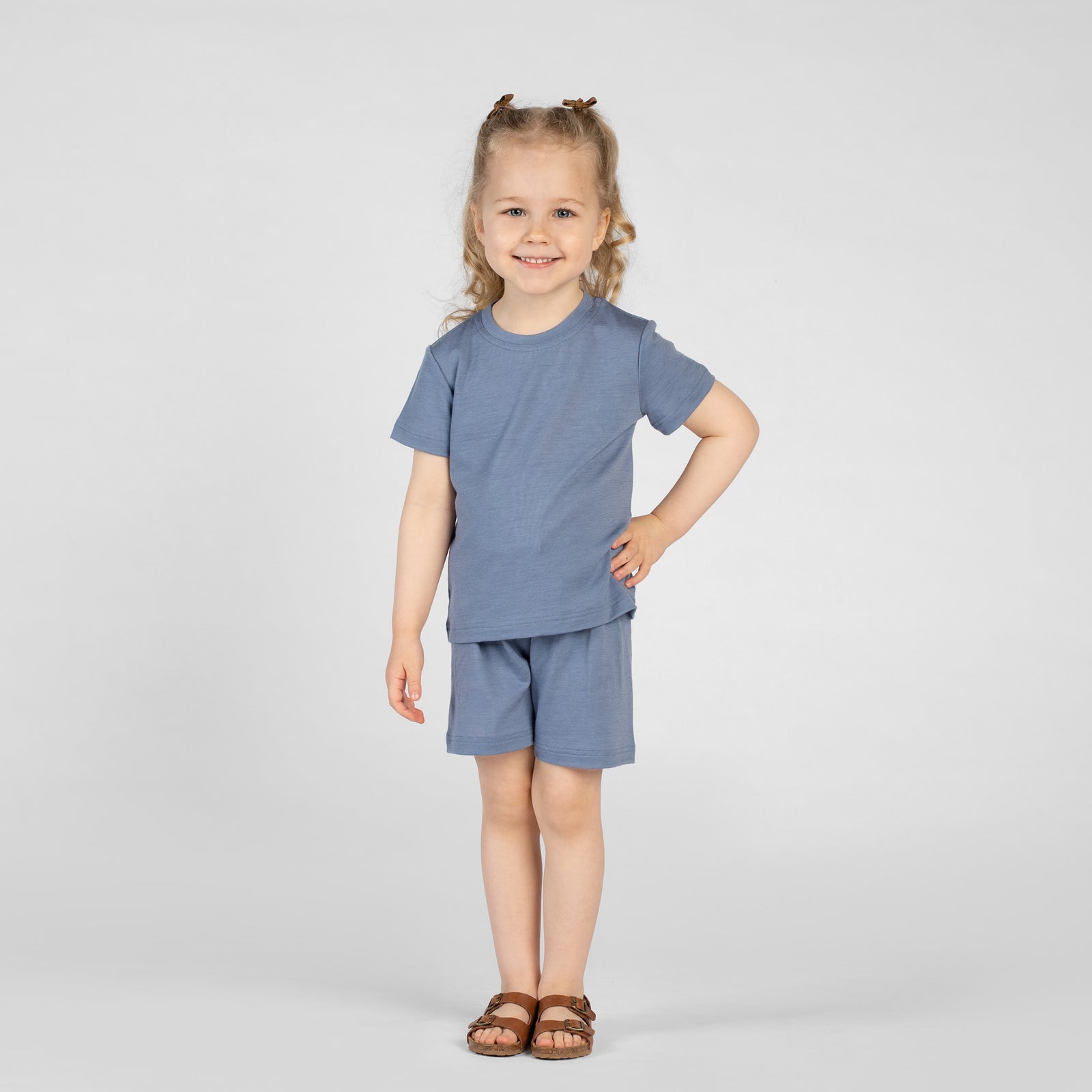 A young child poses playfully with one hand on their hip and a bright smile, wearing a dusty blue short-sleeve t-shirt and matching shorts. They have blonde hair styled in half-up pigtails with small brown clips, set against a plain light background.