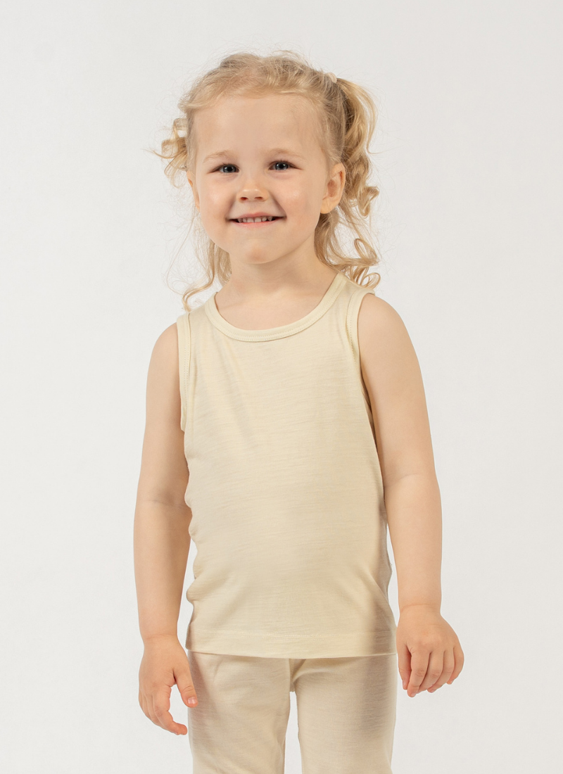 A young girl with curly blonde hair styled in loose pigtails smiles sweetly directly at the camera. She stands against a seamless white background, modeling a light beige scoop-neck tank top and matching bottoms.