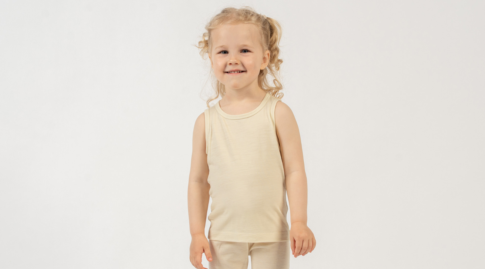 A young girl with curly blonde hair styled in loose pigtails smiles sweetly directly at the camera. She stands against a seamless white background, modeling a light beige scoop-neck tank top and matching bottoms.