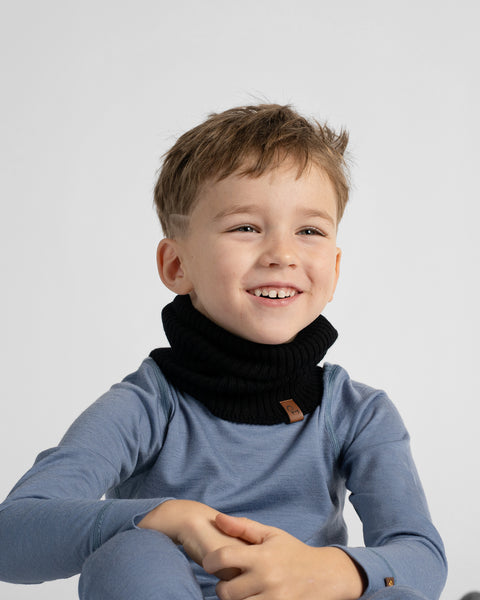 A young boy smiling while wearing a black ribbed knit neck warmer over a dusty blue long-sleeve top, photographed against a clean light gray studio background.