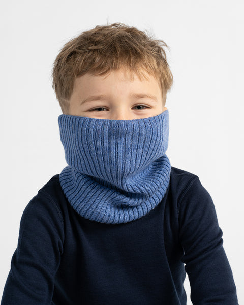 A young child wearing a thick, ribbed knit neck warmer in a dusty medium blue color, styled over a dark navy long-sleeve shirt against a clean white background.
