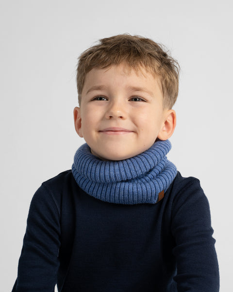 A young boy with short brown hair smiling, wearing a thick blue ribbed knit neck warmer over a dark navy long-sleeve shirt, set against a plain light gray background.