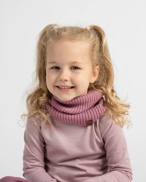 A young girl with blonde pigtails smiling while wearing a dusty pink ribbed knit cowl neck warmer over a matching long-sleeve top, set against a plain light gray background.