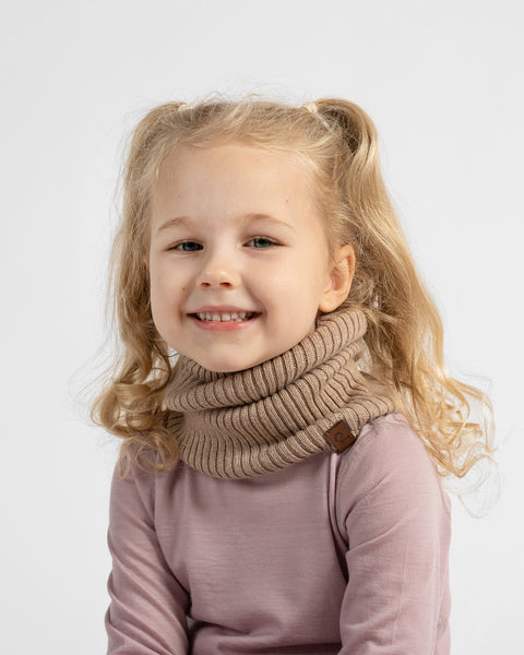 A young girl with blonde pigtails smiling while wearing a beige ribbed knit cowl neck warmer over a mauve long-sleeve top, photographed against a light gray studio background.