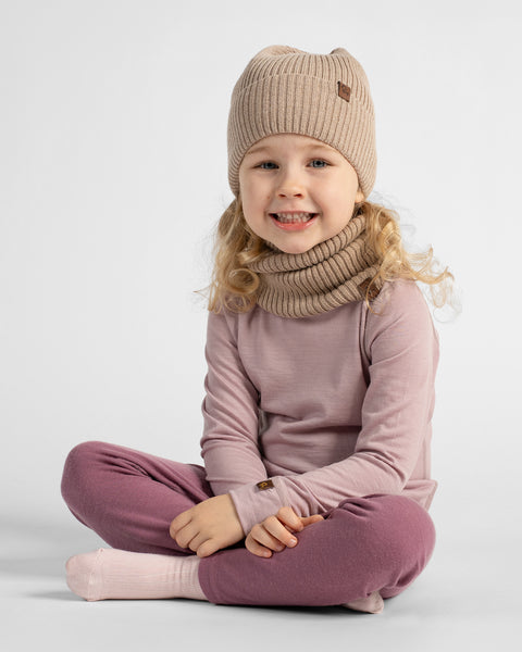 A young child sitting cross-legged wearing a ribbed knit beanie, matching knit neck warmer, mauve long-sleeve top, mauve pants, and pale pink socks against a light gray studio background.