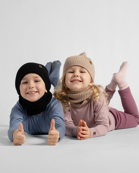 Two children lying on their stomachs and giving thumbs up, wearing matching knit winter hats and neck warmers in a studio setting.