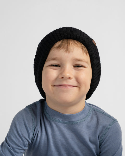 A smiling young boy wearing a black ribbed knit beanie and a dusty blue long-sleeve shirt, photographed against a clean light gray background.